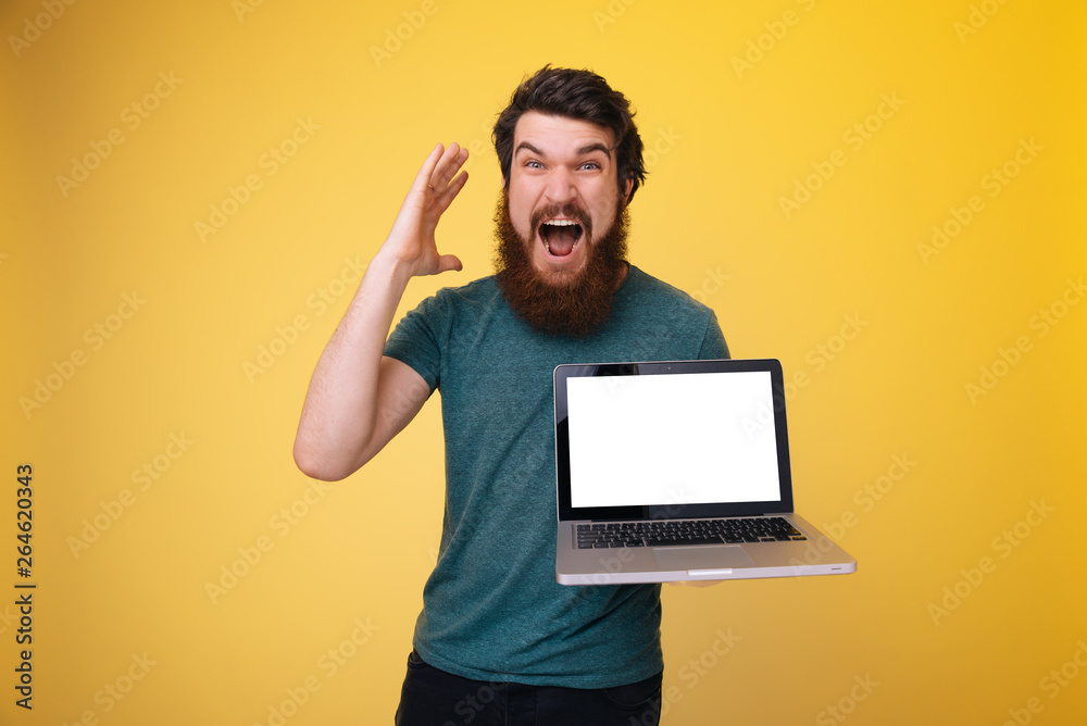 Excited man with beard is olding a laptop, an looking amazed at camera over yellow background