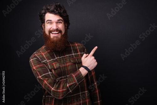 Portrait of bearded man looking at camera while poiting away with finger over darg isolated background