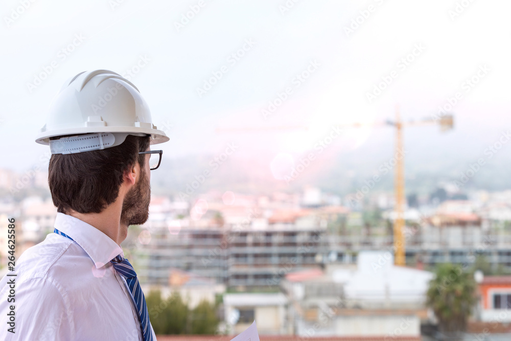Giovane ingegnere edile con occhiali da vista, caschetto e camicia bianco, cravatta blu. Di fronte a un cantiere edile con gru e palazzina in costruzione visto di spalle. Giovane ingegnere edile con occhiali da vista, caschetto e camicia bianco, cravatta blu. Di fronte a un cantiere edile con gru e palazzina in costruzione visto di spalle.