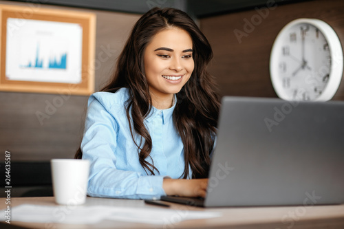 Photo of beautiful young manager woman with bright smile working with laptop in modern office.