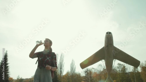 Wide shot of young attractive girl in a red plaid shirt drinkig water from a plastic bottle. Vintage plane on a background photo