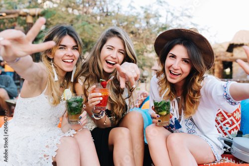 Canvas Print Attractive long-haired girls in stylish attires and accessories drink cocktails and posing with hands waving