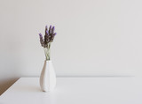 Close up of lavender sprigs in small white vase on side table against neutral wall background (selective focus)