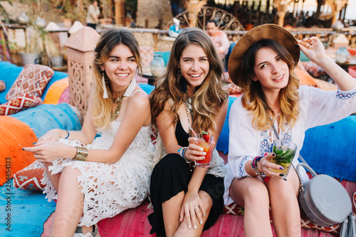 Portrait of three graceful sisters in vintage outfit resting on blue sofa with glasses of soda and cocktail. Cheerful girls having fun outside and gladly posing with sincere smile, waiting for friends