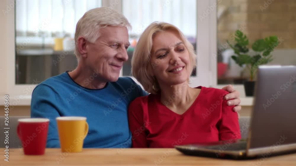 Closeup shoot of senior happy couple using the laptop on the desk with cups with tea smiling happily indoors in a cozy apartment Closeup shoot of senior happy couple using the laptop on the desk with cups with tea smiling happily indoors in a cozy apartment