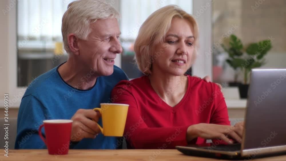 Closeup shoot of senior happy couple networking on the laptop on the desk and drinking coffee indoors in a cozy apartment Closeup shoot of senior happy couple networking on the laptop on the desk and drinking coffee indoors in a cozy apartment