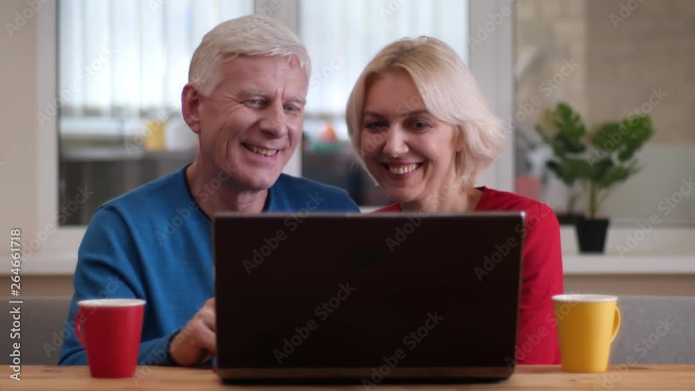Closeup shoot of aged happy couple having a video call on the laptop with cups with tea on the desk indoors in a cozy apartment Closeup shoot of aged happy couple having a video call on the laptop with cups with tea on the desk indoors in a cozy apartment