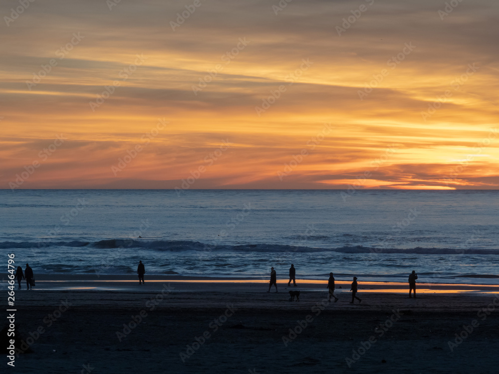 Fototapeta premium Beach visitors walking along Pacific Ocean with sun setting during dusk 