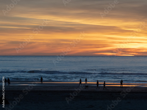 Beach visitors walking along Pacific Ocean with sun setting during dusk 