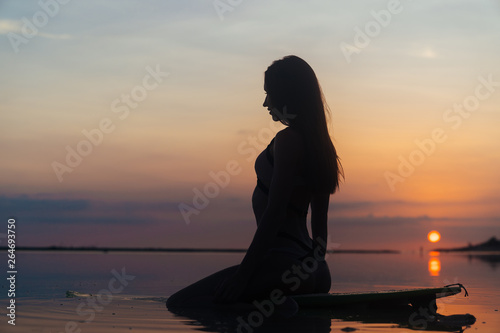 Silhouette sexy girl in swimsuit lying and posing on surf board at beach during sunset
