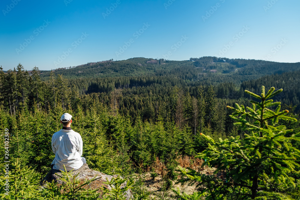 Young man sit on rock with forest and blue sky, looking to valley
