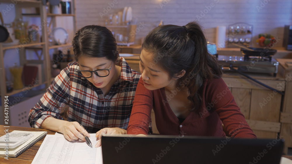 young asian female roommates in dormitory accounting math on calculator working on expense reading document paper using laptop computer and calculate. woman in glasses explain to friend in kitchen. young asian female roommates in dormitory accounting math on calculator working on expense reading document paper using laptop computer and calculate. woman in glasses explain to friend in kitchen.