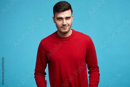 Handsome young dark haired male wearing stylish red sweater with long sleeves smiling at camera, posing against blue studio wall background with copyspace for your text or advertising content