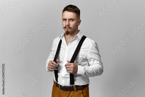 Studio picture of attractive elegant young groom with handlebar mustache and beard dressing up, wearing white shirt and brown trousers, adjusting suspenders, looking at camera with serious expressin photo