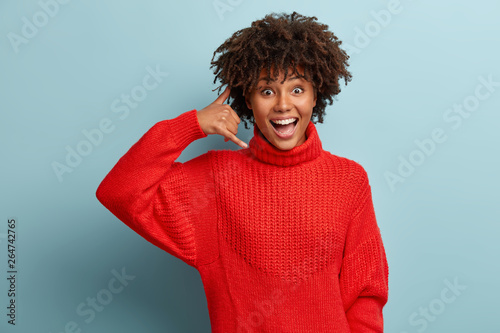 Konstfotografi Happy overjoyed Afro American woman shows call me gesture, asks keep in touch, wears warm red sweater, being in high spirit, isolated over blue studio background