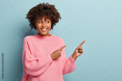 Portrait of happy delighted woman points in right direction on copy space, gazes happily, advertizes nice product, wears oversized pink jumper, isolated on blue wall. Check out cool advert there photo