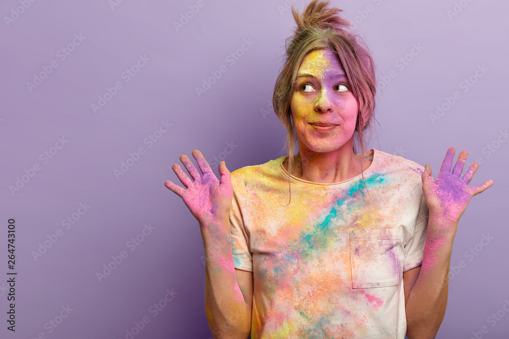 Carefree beautiful female model looks aside with dreamy curious expression, raises palms smeared with colorful powder, involved in Holi festival celebrations, stands over purple wall, blank space Carefree beautiful female model looks aside with dreamy curious expression, raises palms smeared with colorful powder, involved in Holi festival celebrations, stands over purple wall, blank space
