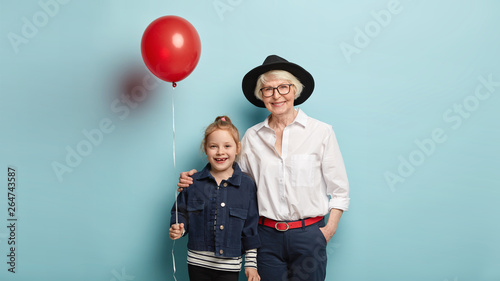 Family, leisure, joy, lifestyle, relationship concept. Positive little kid holds air balloon, celebrates holiday together with affectionate grandma in stylish clothes. Two generations on photo. photo