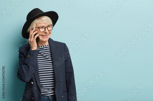 Beautiful grey haired lady has telephone conversation after working day, calls via application, discusses new succesfully completed task, wears stylish headgear, isolated over blue background photo