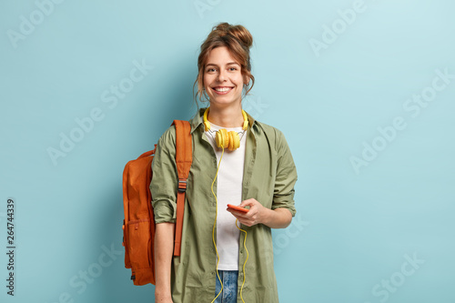 Beautiful smilig female traveller has leisure time, enjoys online communication, connected to headphones, listens music from playlist, wears casual white t shirt and green shirt, carries rucksack photo