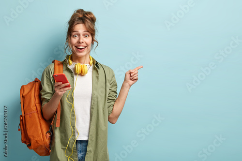 Studio shot of happy teenage girl holds mobile phone, checks email, gets new notification, uses modern headphones, points at copy space for brand name or label, wears loose khaki shirt, carries bag photo