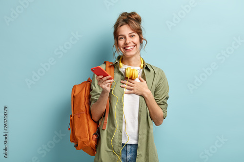 Indoor shot of pleased European student happy to receive message of congratulation after passing exam, holds palm on chest, demonstrates gratitude, listens music with headphones, wears stylish clothes