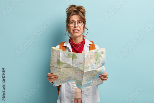 Crying female tourist lost in unknown destination, holds map, tries to find way, looks with dissatisfied dejected expression, wears round spectacles, has rucksack with packed things, stands indoor photo
