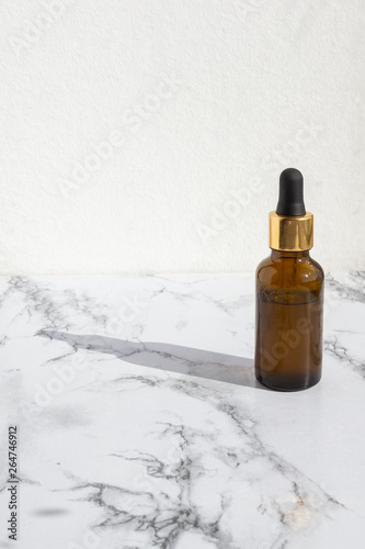 Pipette dropper with drop of natural oil above brown glass bottle on marble background. Natural organic skin care beauty products.