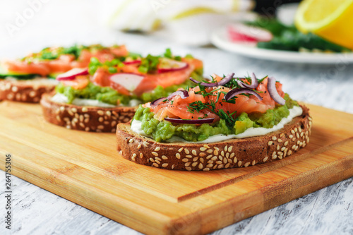 three open sandwiches with toast bread smoked salmon cream cheese sliced cucumber dill avocado red onion on wooden cutting board close up soft focus
