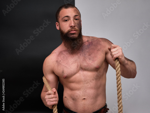 Young brutal man with a beard, bodybuilder with a naked torso posing, talking on a black and white background in the studio with a rope. Beautiful male body. Concept art.