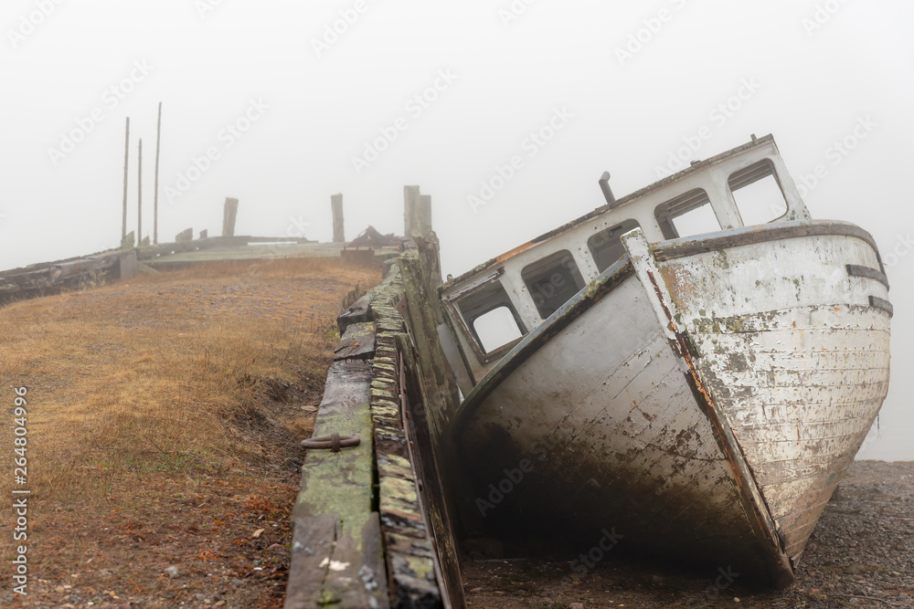 A wrecked boat on the shore next to an old abandoned wharf on a very foggy day. The boat is in very poor shape and is falling apart. Front view at low tide.
