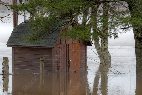 A small shack under trees surrounded by flood water. Foggy river in the background. The water is about one third of the way up the building.