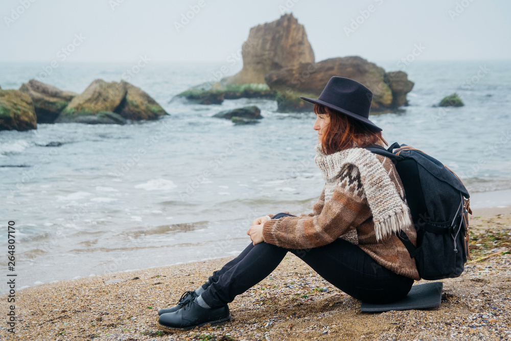 Young woman tourist in hat and with backpack sitting on beach, looking at sea, on coastline, on horizon. Tourism, vacation, lifestyle. Hipster girl resting on beach