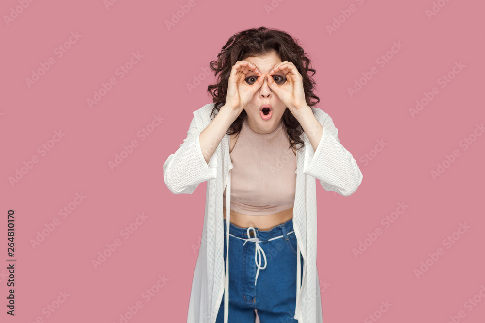 Portrait of shocked brunette young woman with curly hairstyle in casual style standing with binoculars hands gesture on big eyes and looking at camera. indoor studio shot isolated on pink background.