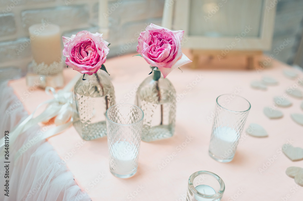 Large pink roses stand in glass vases along with candles in glass cups. Wedding ceremony. Scenery and details. Evening rites and traditions. Large pink roses stand in glass vases along with candles in glass cups. Wedding ceremony. Scenery and details. Evening rites and traditions.