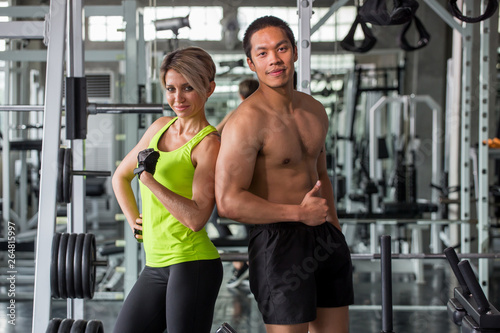 portrait of couple in sportswear posing strong showing her biceps muscular arms and Thumbs up in fitness gym . sport asia man and caucasian woman workout ,training.