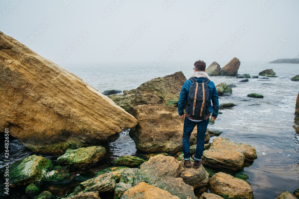 Traveler with a backpack stands on a rock against a beautiful sea with waves, a stylish hipster boy posing near a calm ocean during a wonderful journey around the world. Shoot from the back