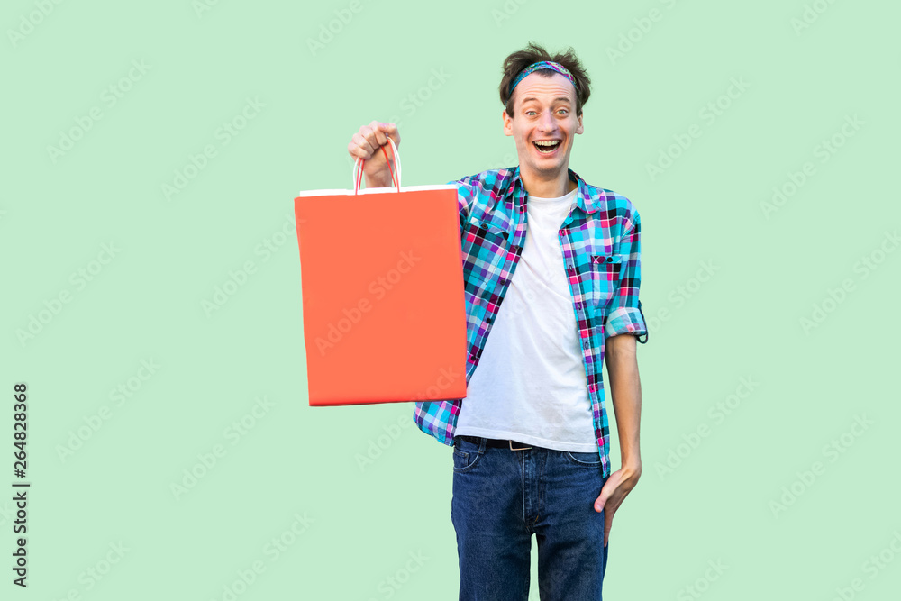 Wow! Shocked modern young hipster man in white t-shirt and checkered shirt standing and showing shopping bags and toothy smile, looking at camera. Studio shot, green background, isolated, indoor Wow! Shocked modern young hipster man in white t-shirt and checkered shirt standing and showing shopping bags and toothy smile, looking at camera. Studio shot, green background, isolated, indoor