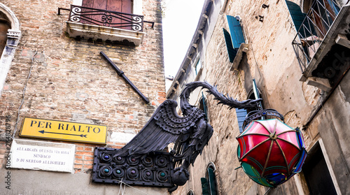Iron dragon hanging from a historic building in Venice, Italy (road sign 'per Rialto means 'to Rialto; white plate translation: 'this road was widened in 1884 Mayor D. Di Serego Allighieri') photo