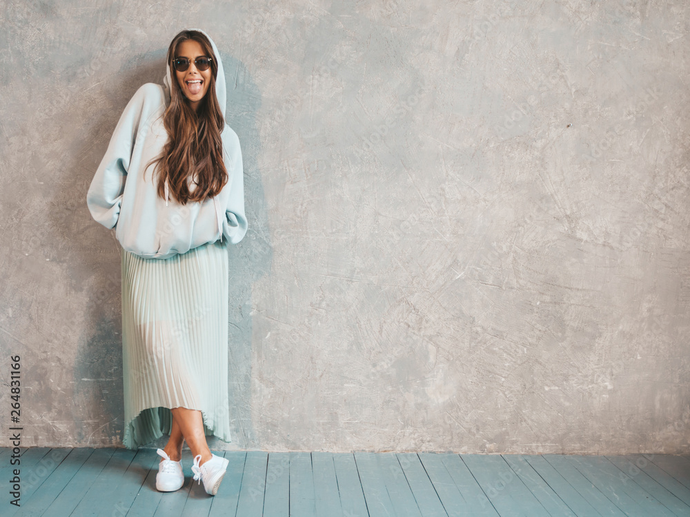 Young beautiful smiling woman looking at camera. Trendy girl in casual summer hoodie and skirt clothes. Funny and positive female posing near gray wall in studio. In sunglasses showing tongue Young beautiful smiling woman looking at camera. Trendy girl in casual summer hoodie and skirt clothes. Funny and positive female posing near gray wall in studio. In sunglasses showing tongue