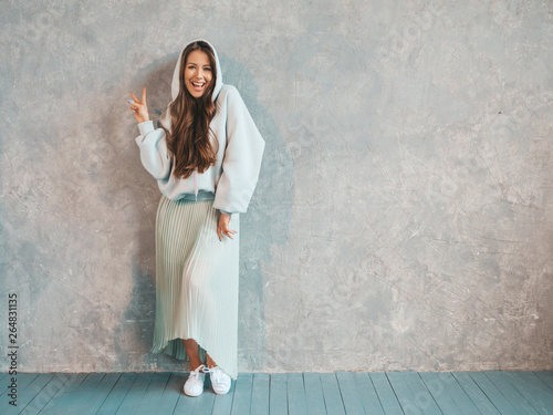 Young beautiful smiling woman looking at camera. Trendy girl in casual summer hoodie and skirt clothes. Funny and positive female posing near gray wall in studio. Showing peace sign photo