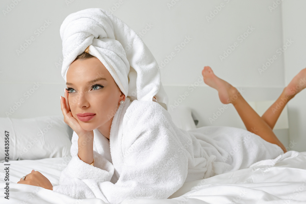 A young girl with white hair in her bedroom is posing on a bed in a white coat and a white towel on her head. A young girl with white hair in her bedroom is posing on a bed in a white coat and a white towel on her head.