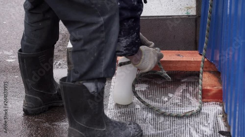 Male worker wearing black rubber boots ties up white plastic bottle with metallic spray tip with thick rope standing outside in street on cold snowing day closely to facade building. photo