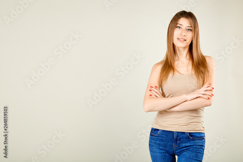 girl in a beige t-shirt and blue jeans is standing against the gray wall is looking at the camera with her arms crossed