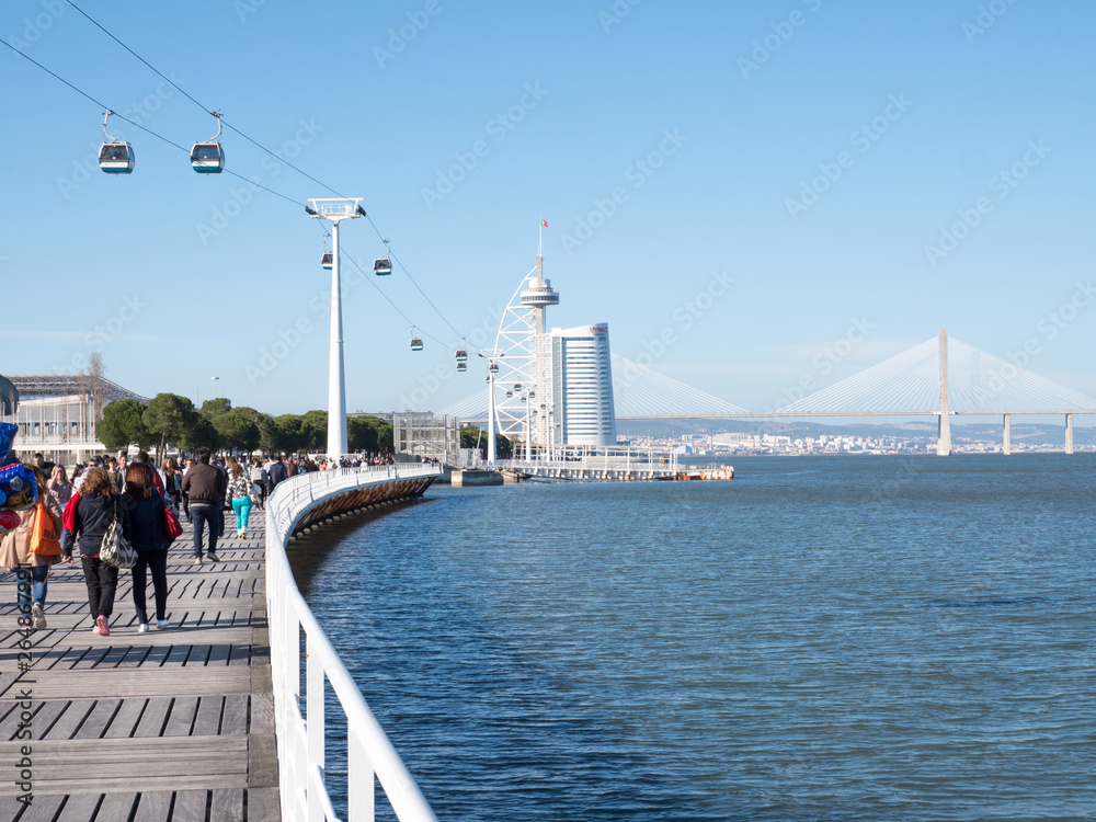 People wakling on a seafront parapet and looking to a sea and bridge in Lisbon