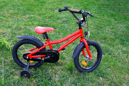Red bike for children with extra side wheels standing on green grass outdoors in park or yard in spring day. Bicycle sport activity equipment for kids.