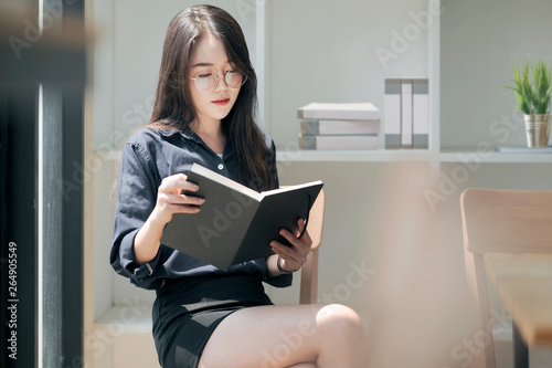 Young asian woman reading a book while sitting near window in white room.