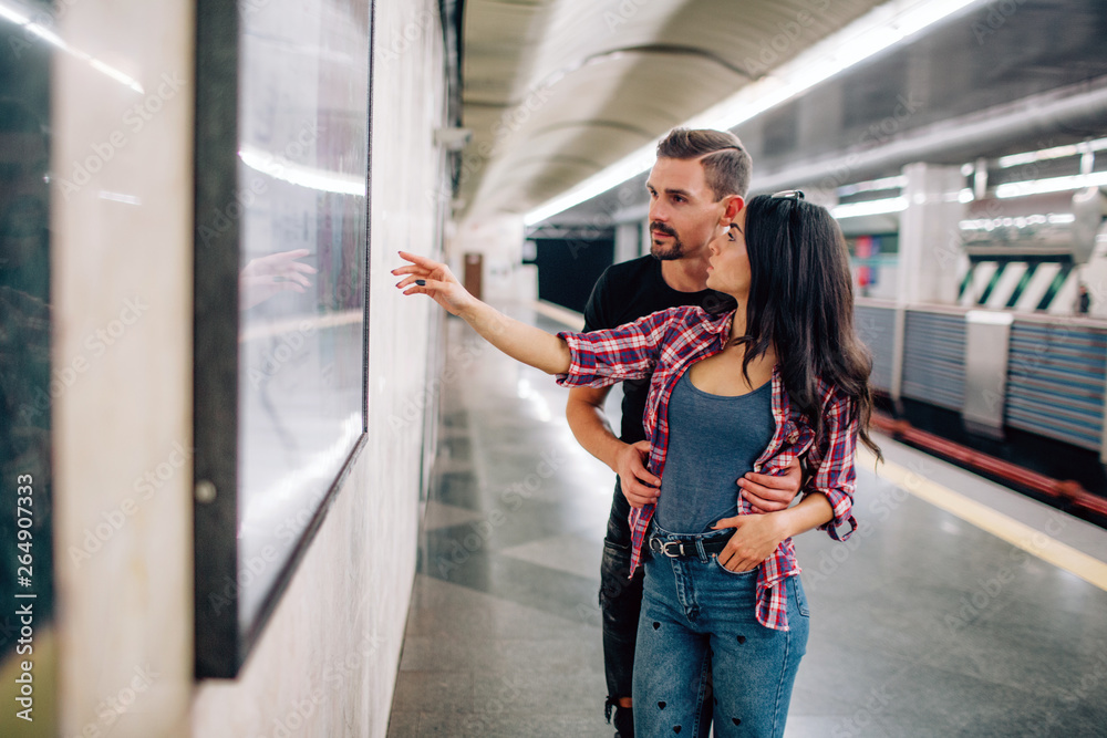 Fototapeta premium Young man and woman use underground. Couple in subway. Casual people in metropolitan. Young woman point on wall and smile. Guy hold hands on her hips.