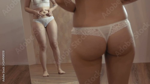 A young girl in lingerie is looking at her body in the mirror. Mild obesity. It touches fat on the body. Static camera.