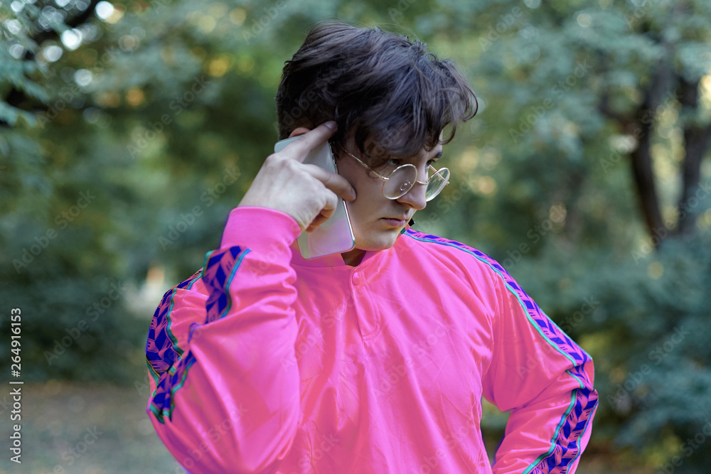 Young handsome caucasian man talks on mobile phone standing in the park. Round golden glasses, bright lilac shirt, extravagant look. Outdoors, copy space. Young handsome caucasian man talks on mobile phone standing in the park. Round golden glasses, bright lilac shirt, extravagant look. Outdoors, copy space.